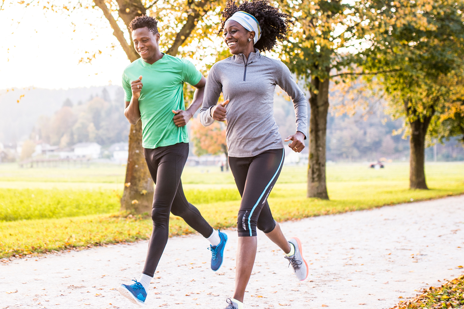  A man and a woman jog side by side on a park's dirt path, smiling at each other.